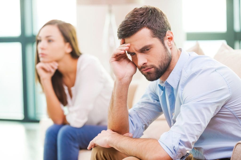 Young man holding hand on head looking away while woman sitting behind him on the couch